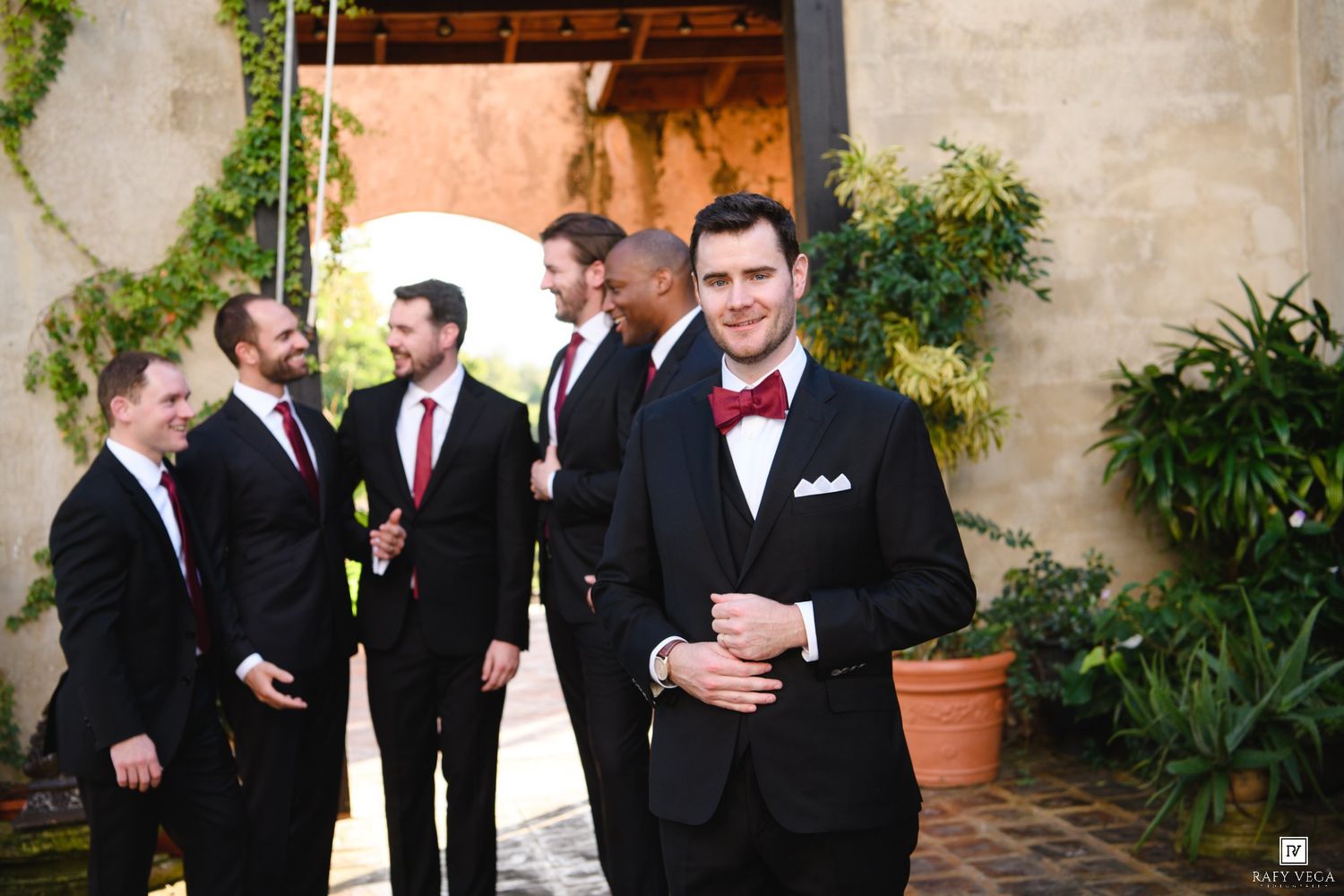 Groom in black tuxedo with red bow tie poses with groomsmen in matching suits at outdoor wedding venue.