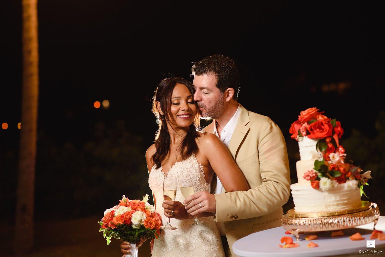 Bride and groom celebrate with champagne beside their floral wedding cake at an outdoor evening reception.