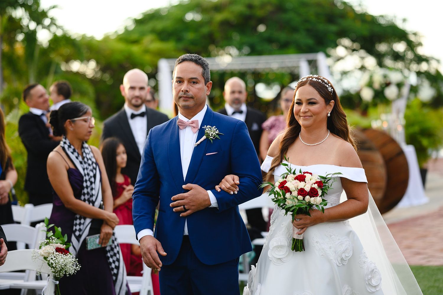 Bride in white off-shoulder gown walks with groom in blue suit at outdoor wedding ceremony.