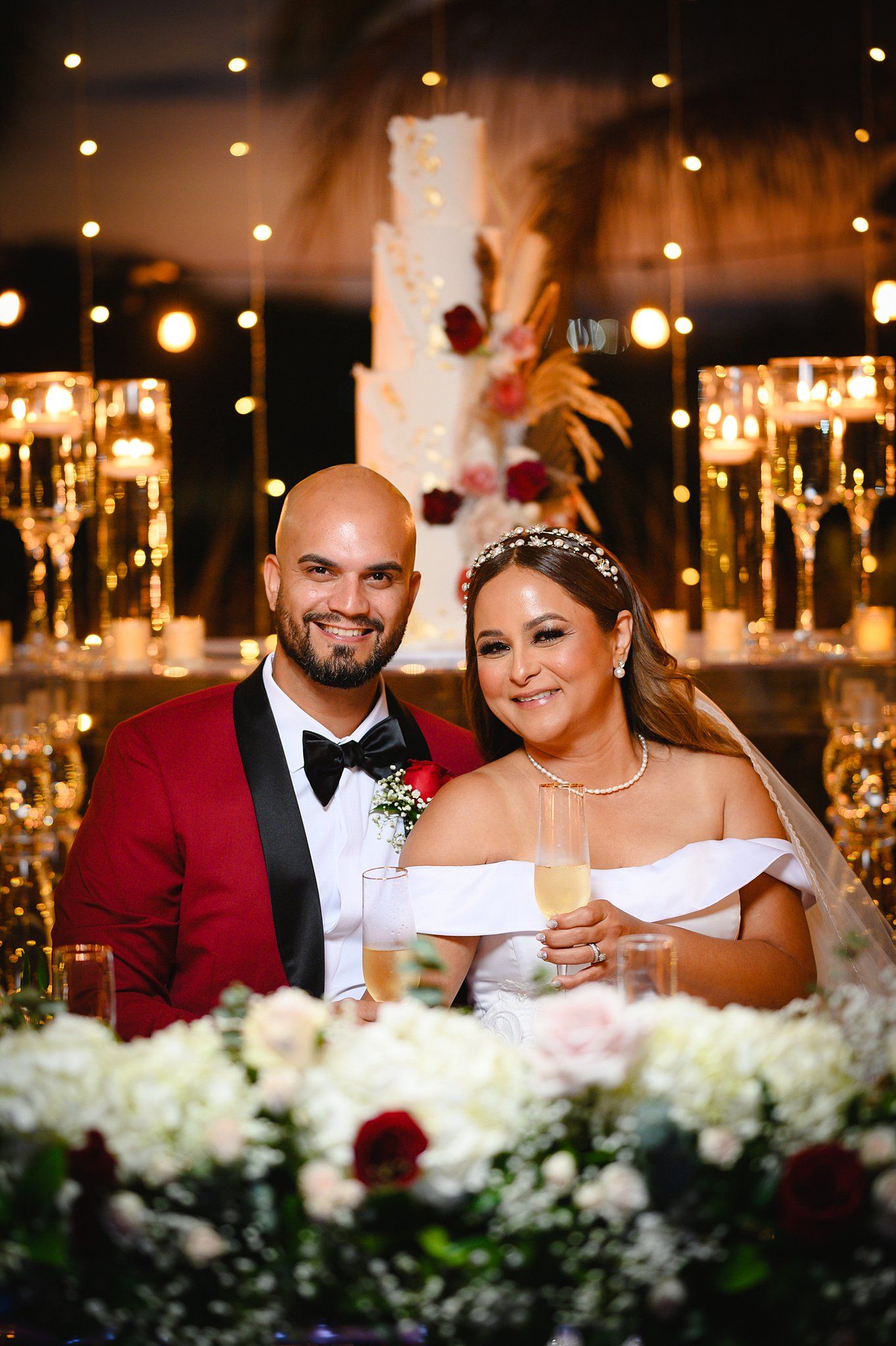 Bride and groom smiling at wedding reception with champagne flutes and elegant white cake backdrop.