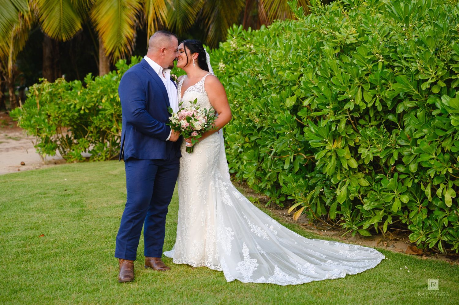 Bride and groom share romantic moment on lush green lawn, bride wearing elegant lace gown with long train.