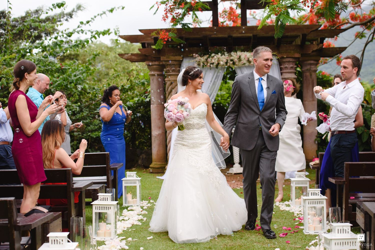 Bride and groom walk down the aisle at an outdoor wedding ceremony while guests celebrate with confetti.