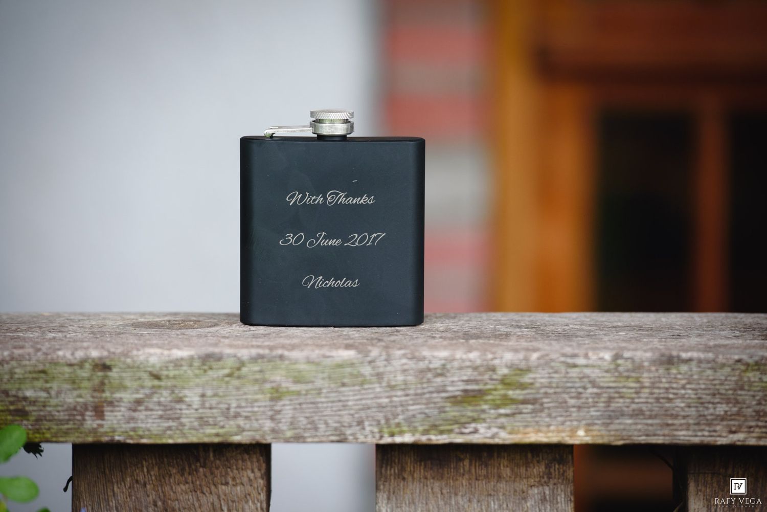 Black engraved hip flask with personalized text, dated 30 June 2017, displayed on a rustic wooden surface.