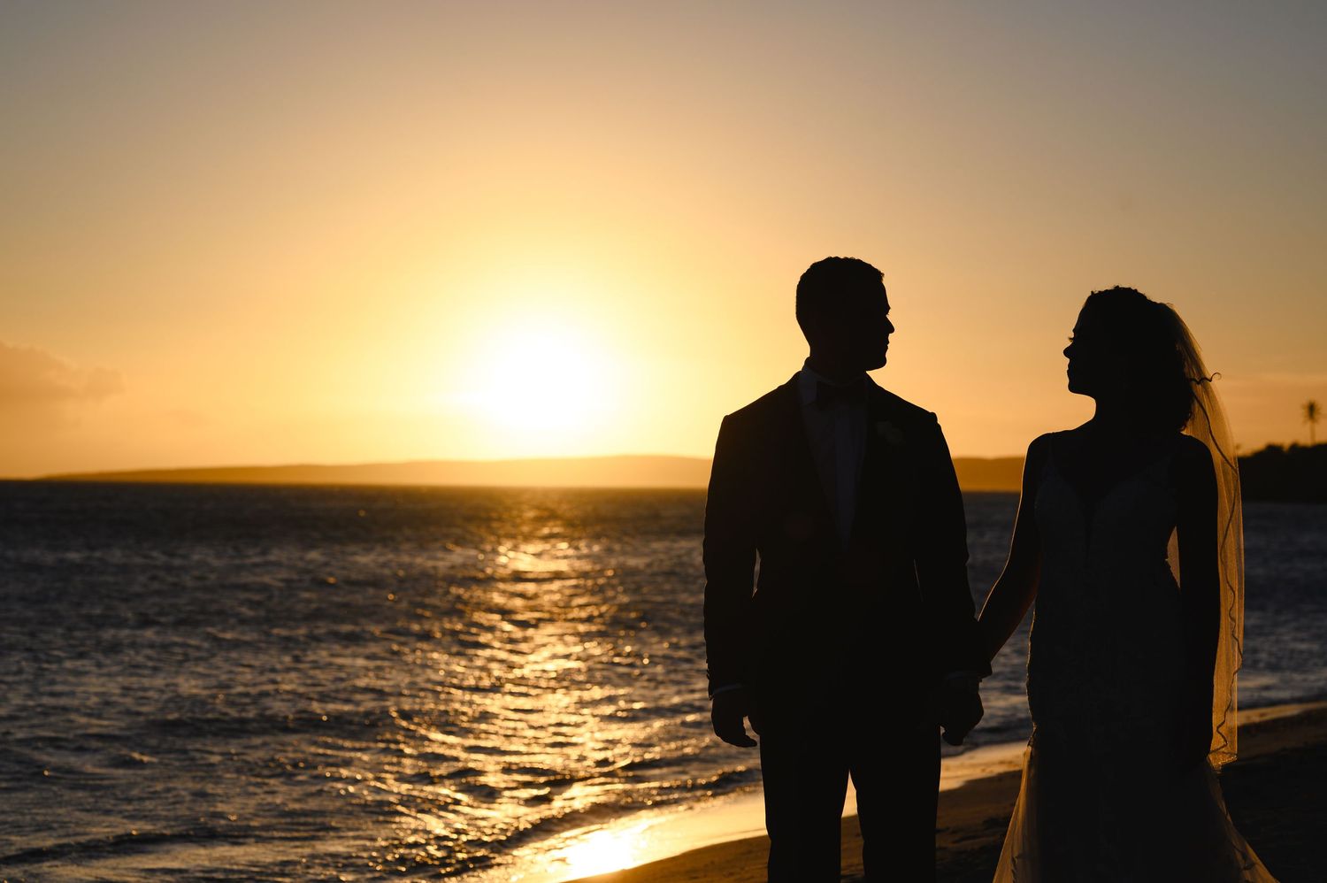 Silhouetted couple in wedding attire holding hands on a beach at sunset, with golden light reflecting on the calm ocean waves.