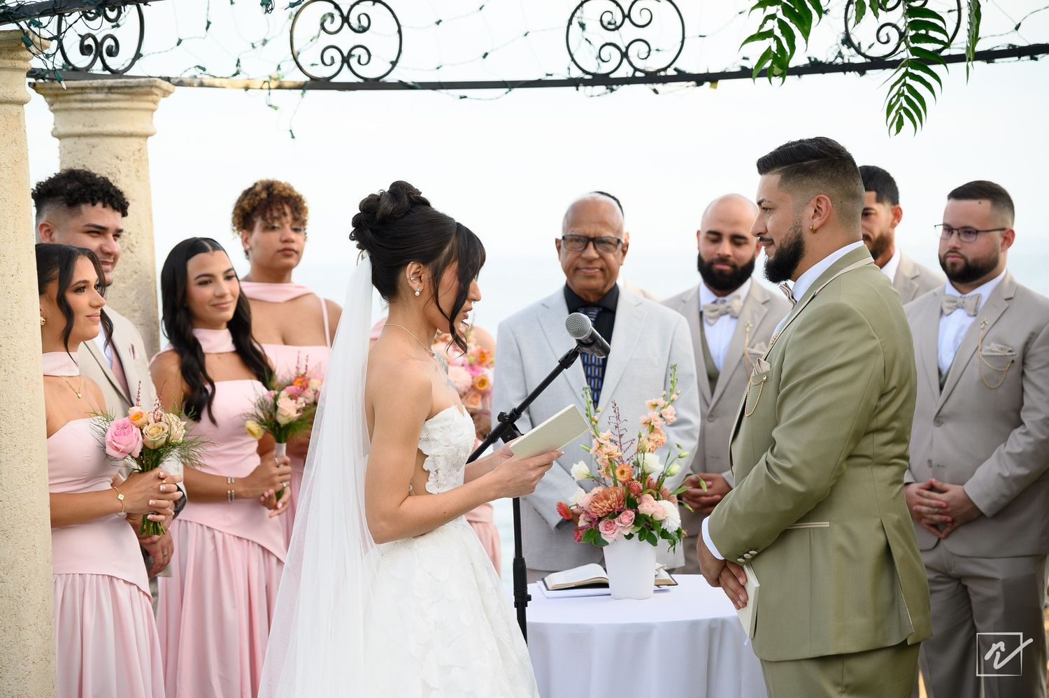 Bride in white gown and groom in olive suit exchange vows at an outdoor wedding ceremony with bridesmaids in pink and groomsmen looking on.