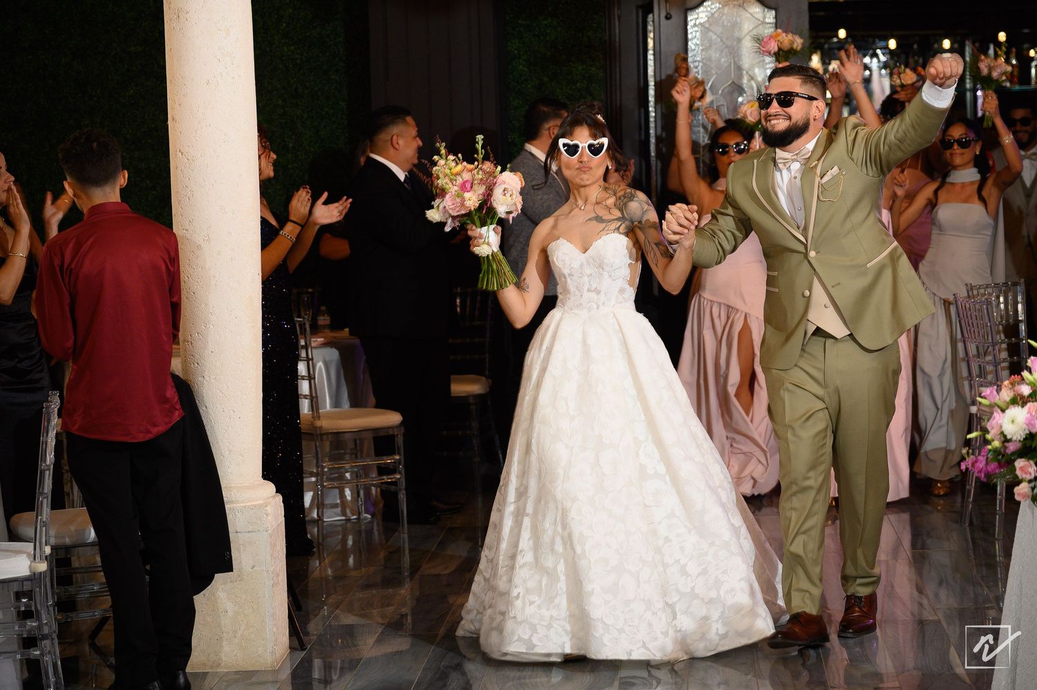 Bride in a lace gown and white sunglasses walks hand in hand with a groom in a green suit as wedding guests cheer.