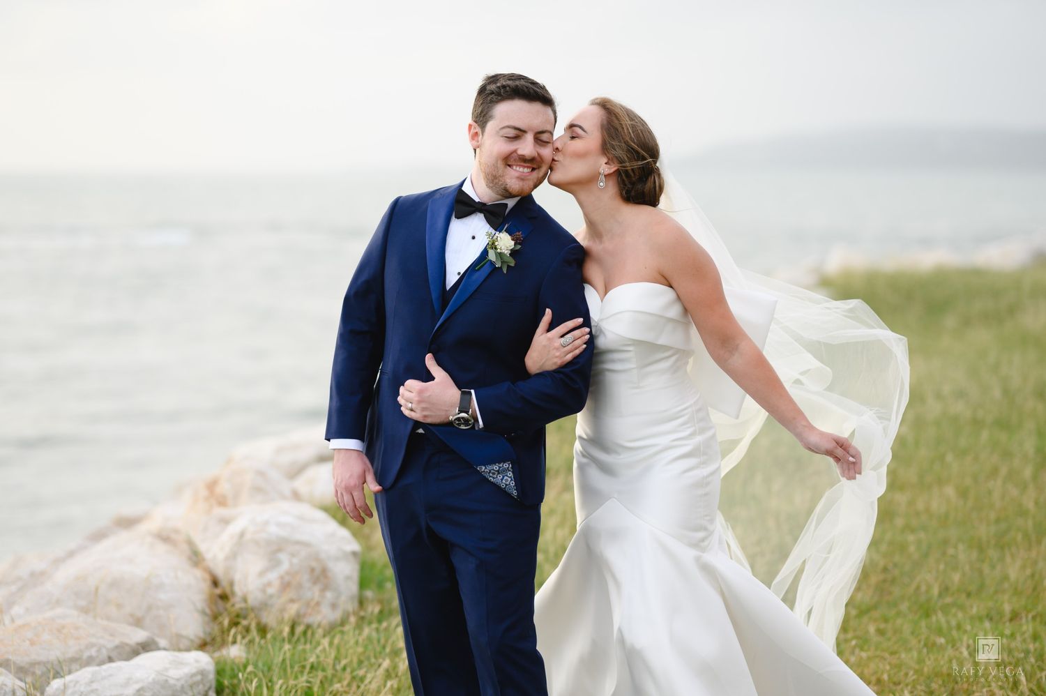 Bride in white strapless gown kisses groom in navy tuxedo outdoors near rocky shoreline with veil flowing in wind.