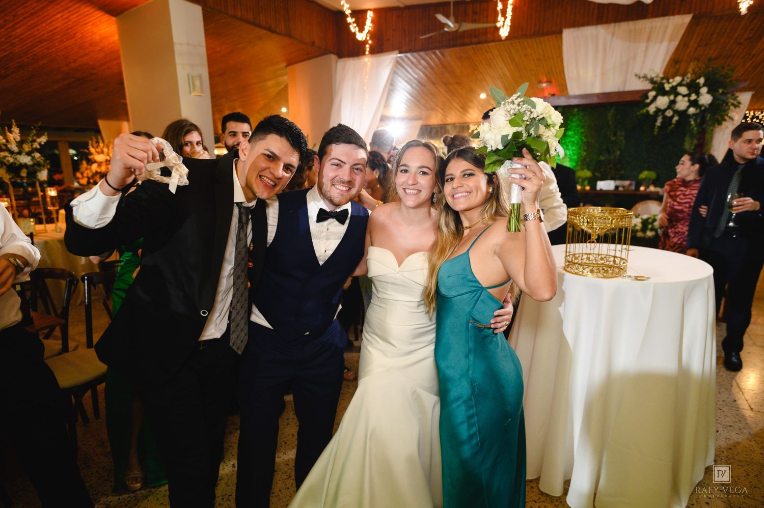 Bride and groom pose with guests at wedding reception, holding bouquet and drinks while smiling joyfully.