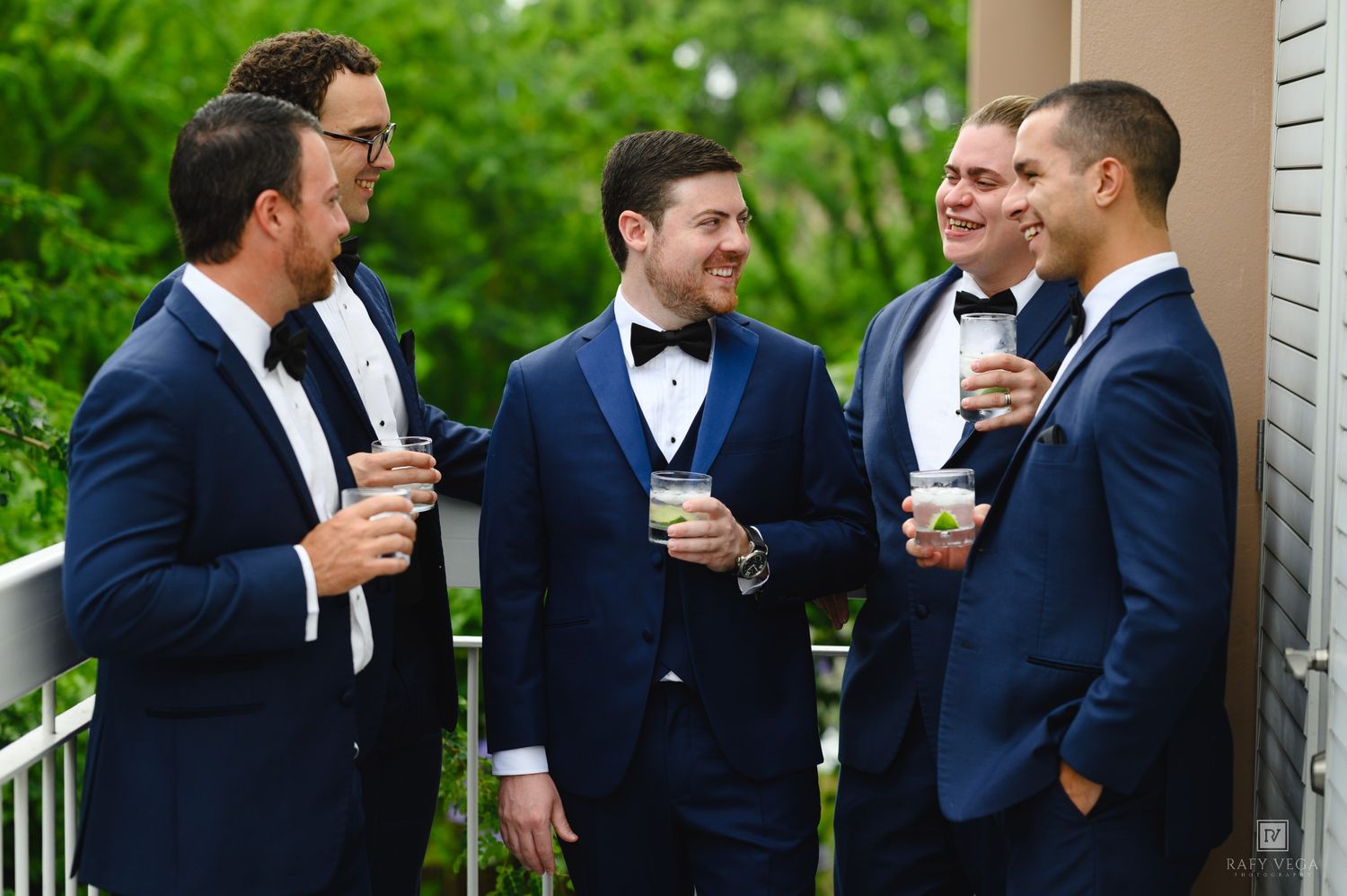 Groom and groomsmen in matching navy blue tuxedos laughing and holding drinks on an outdoor balcony.