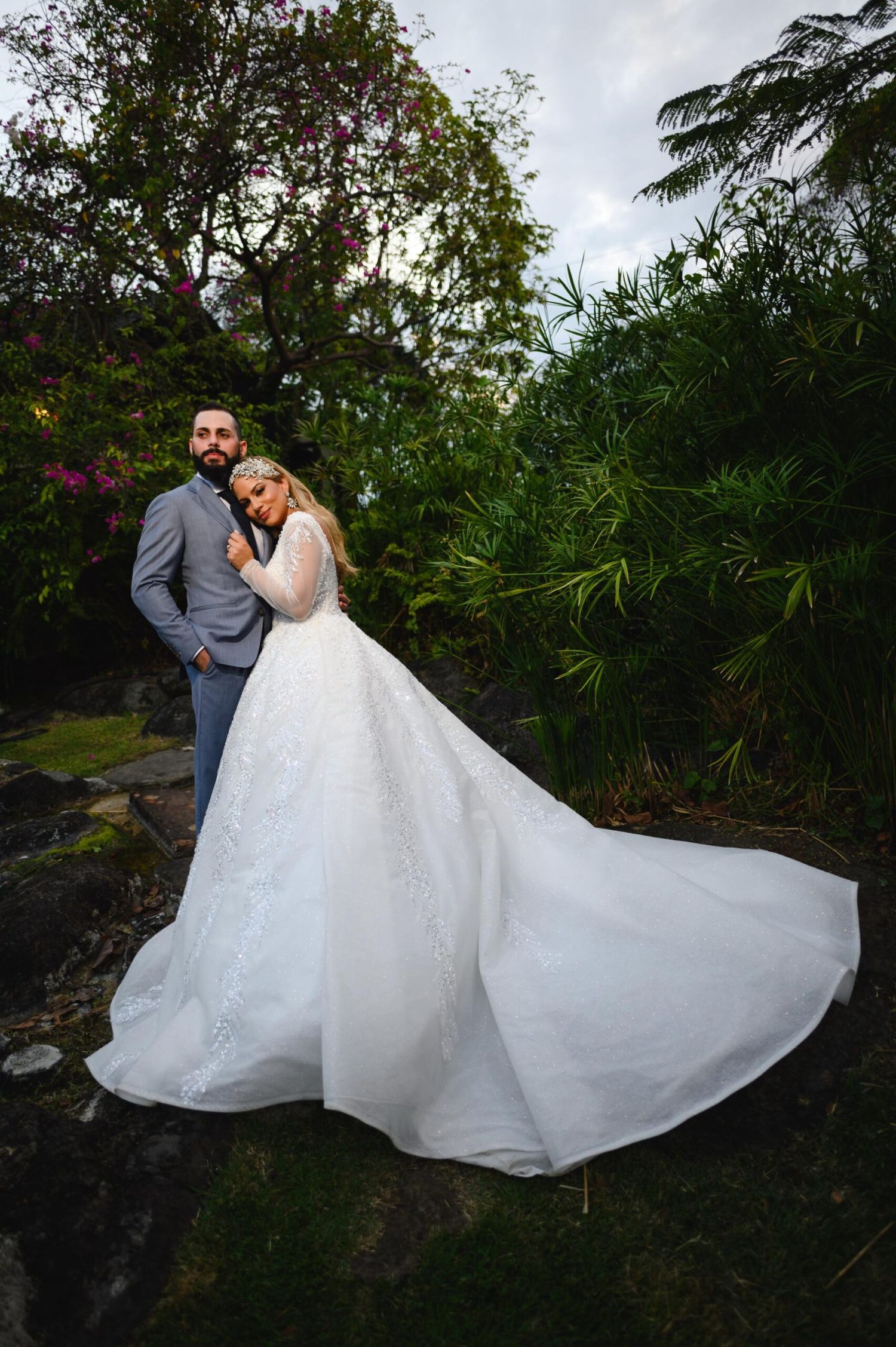 Groom embraces bride in elegant white lace gown with cathedral train amid tropical garden greenery.