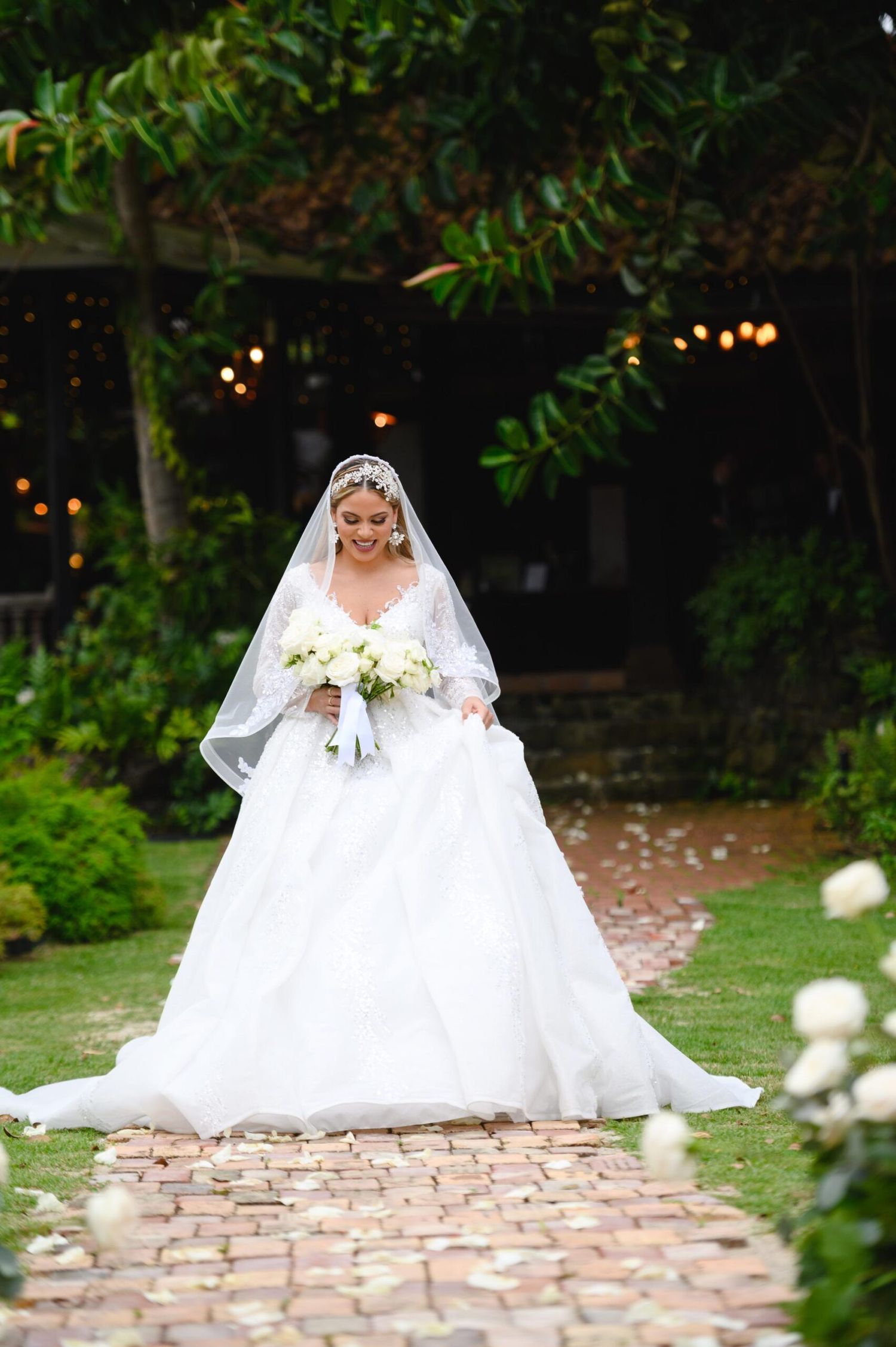 Bride in a white ball gown and veil walks down a brick aisle holding a white bouquet in a lush garden setting.