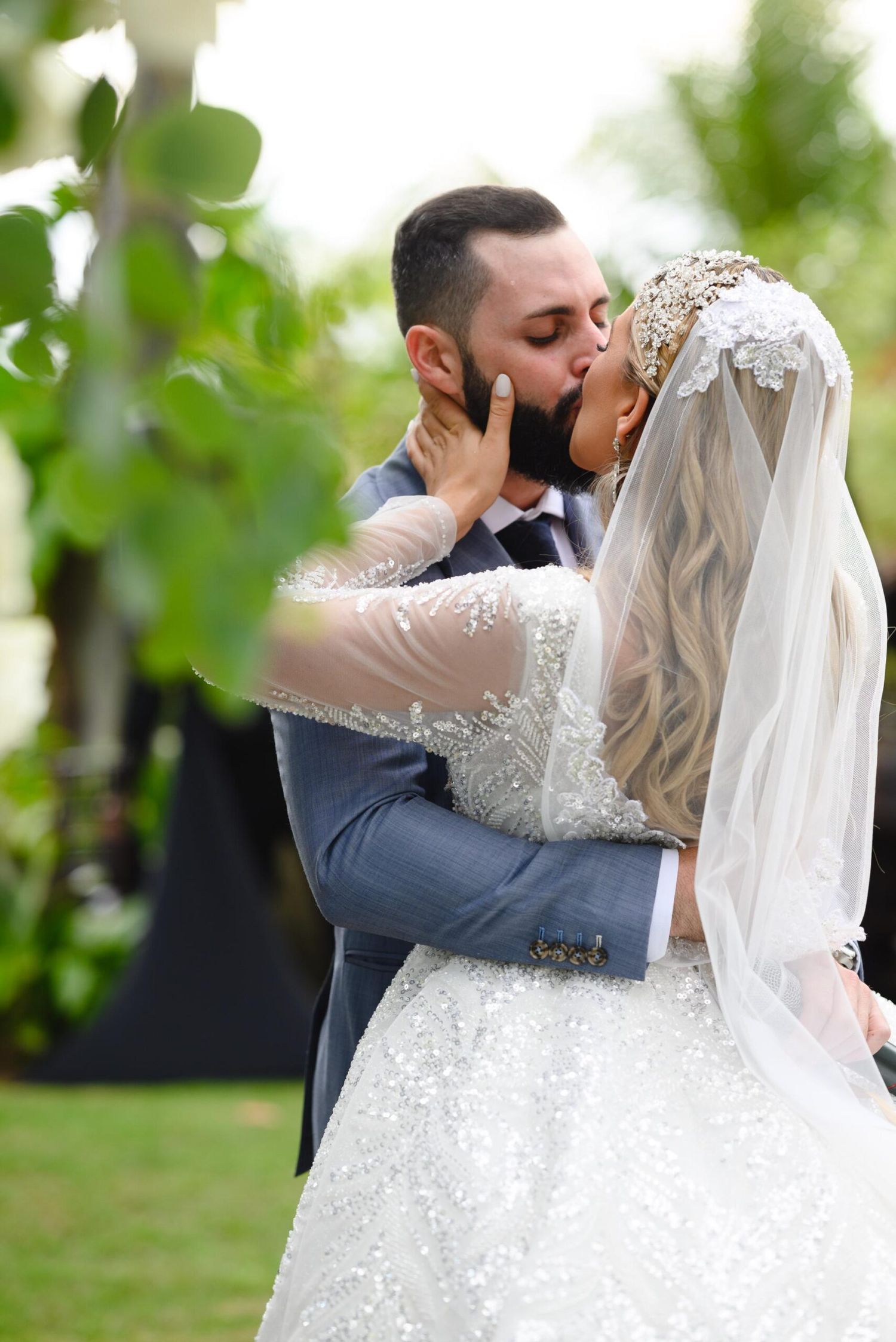 Bride and groom share a romantic first kiss outdoors, bride wearing a beaded gown and veil, groom in a gray suit.