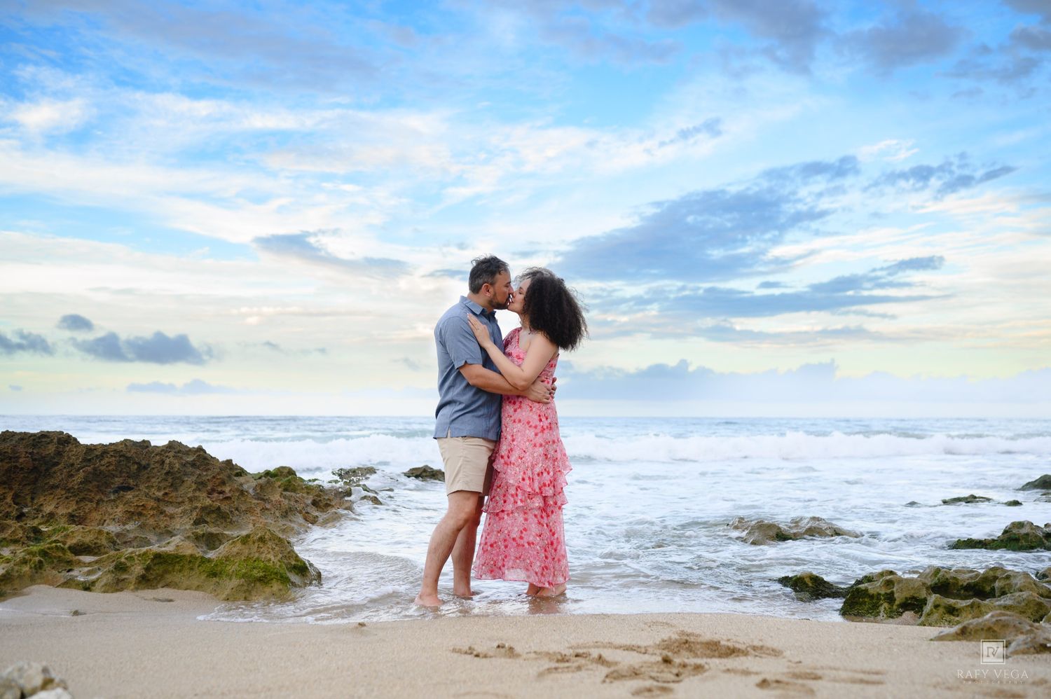 Romantic couple embracing and kissing on a rocky shoreline with a beautiful cloudy sky backdrop.