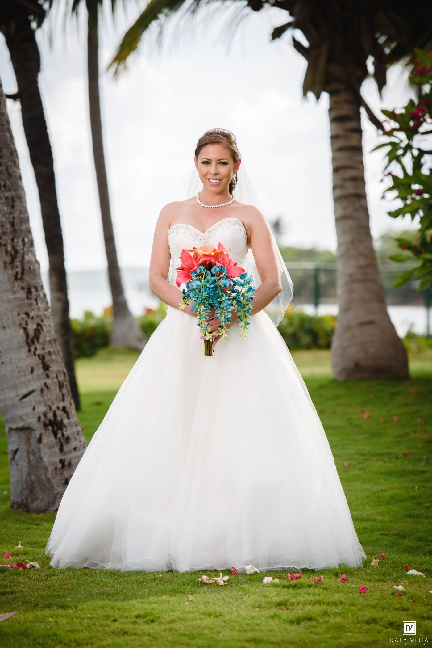 Bride in white ball gown holding vibrant teal and coral bouquet standing on green lawn with palm trees.