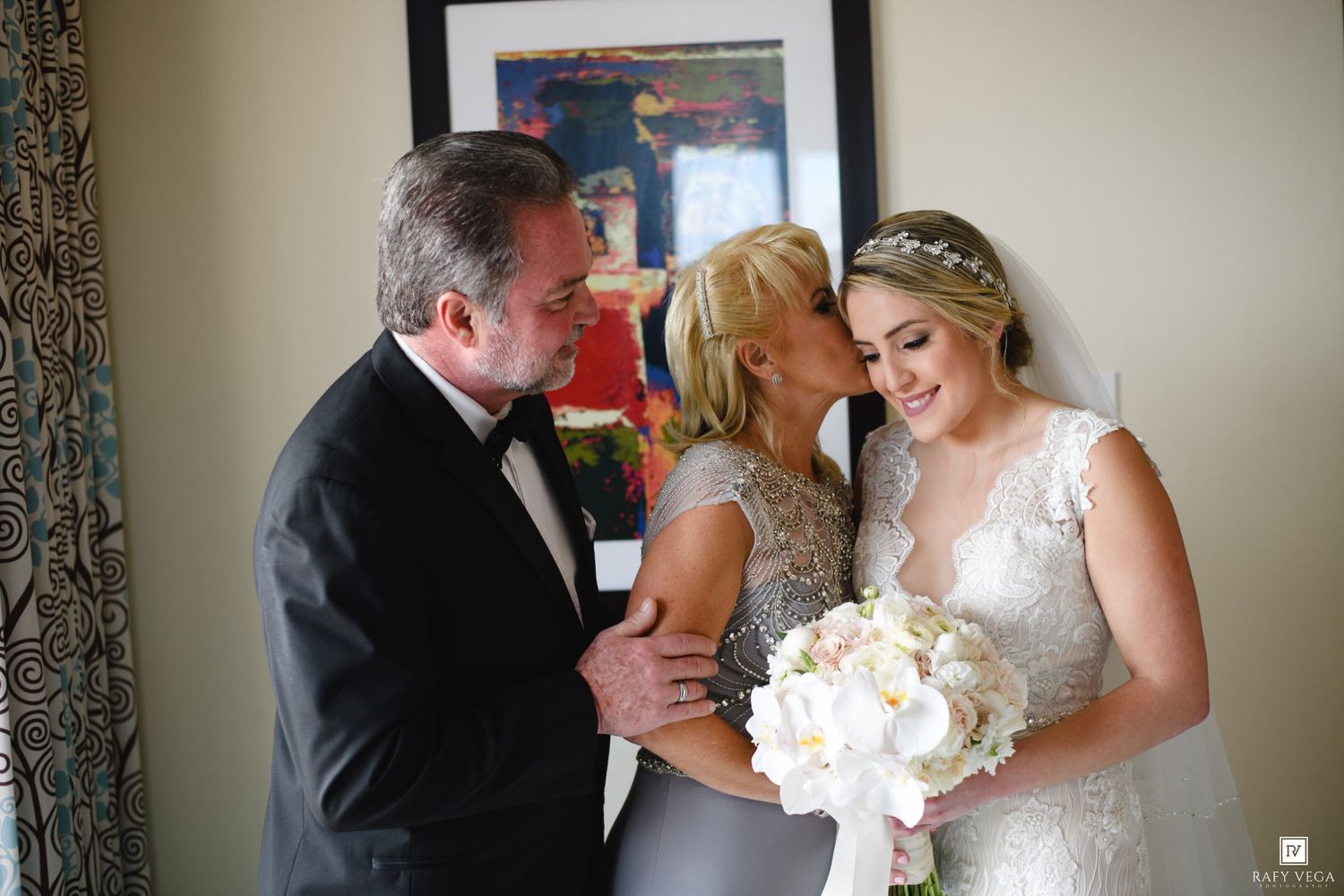 Bride in lace gown holding white bouquet shares tender moment with parents before wedding ceremony.