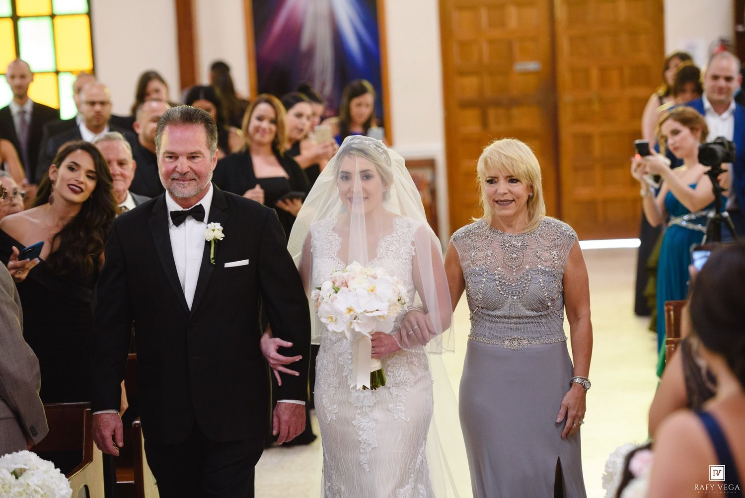 Bride walks down church aisle with parents, wearing white gown and veil, holding bouquet, guests watching ceremony.