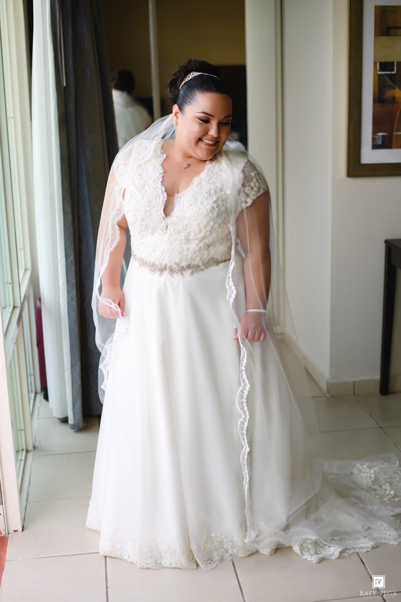 Plus-size bride smiling in white lace wedding gown with veil and beaded belt, standing in bridal suite.