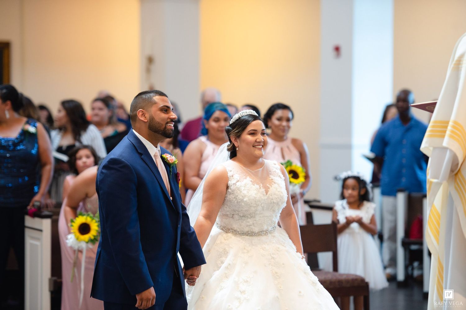 Bride and groom walk down church aisle smiling, bridesmaids with sunflowers and flower girl visible in background.