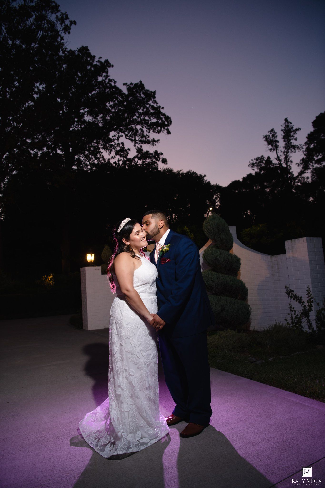 Bride and groom share a romantic kiss at dusk outdoors, bride in white lace gown, groom in navy suit.