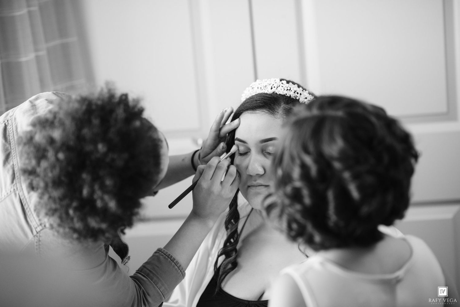 Bride getting makeup applied by two stylists while wearing a tiara on her wedding day.