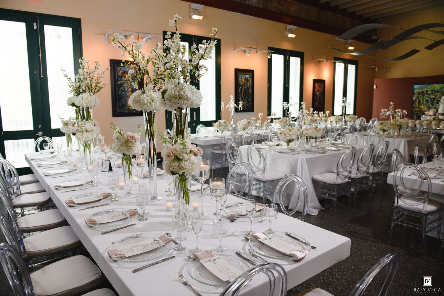Elegant wedding reception hall with white floral centerpieces, long banquet tables, and ghost chairs in a bright venue.