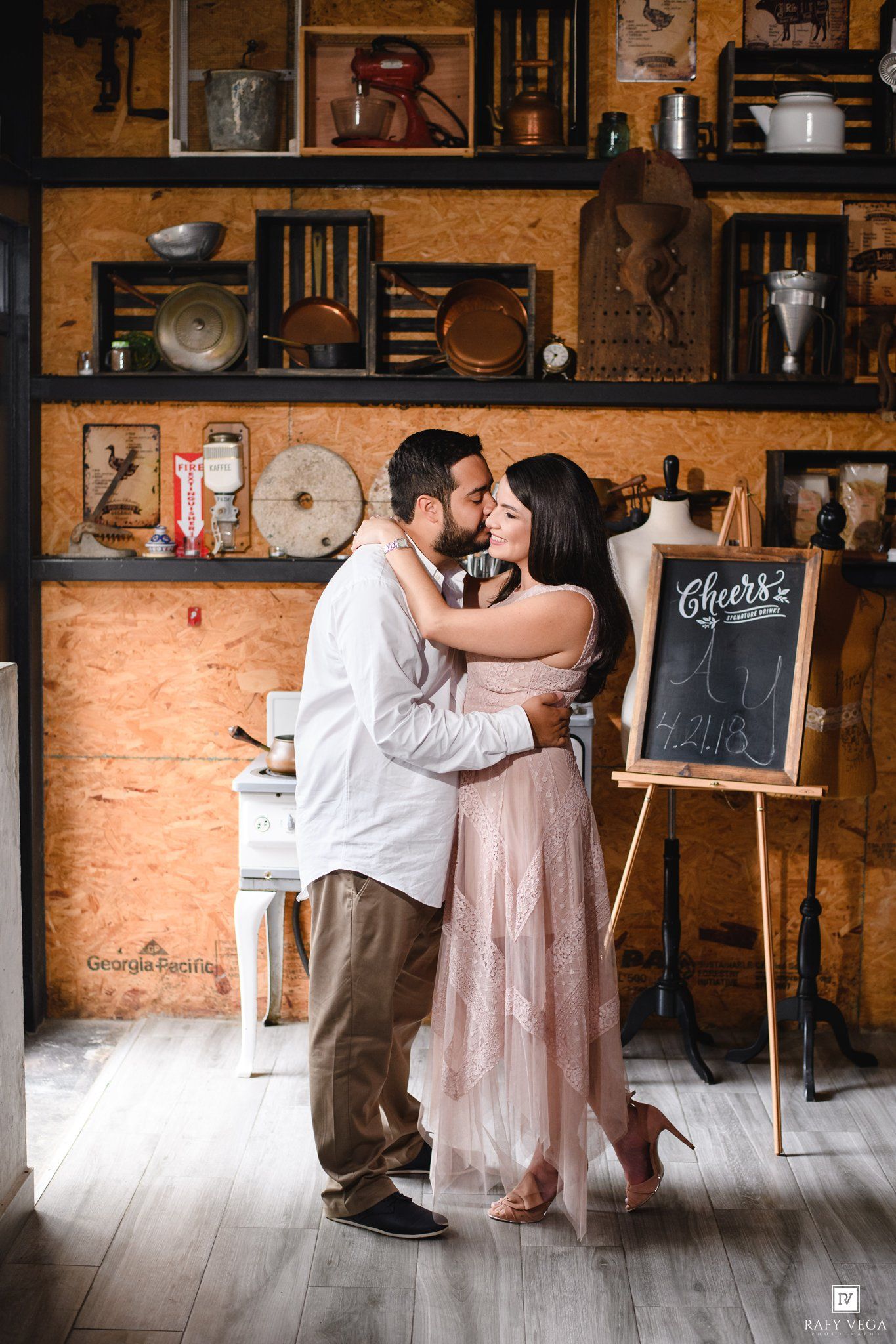 Couple sharing a romantic kiss in a rustic vintage shop with shelves of antiques and a Cheers chalkboard sign.