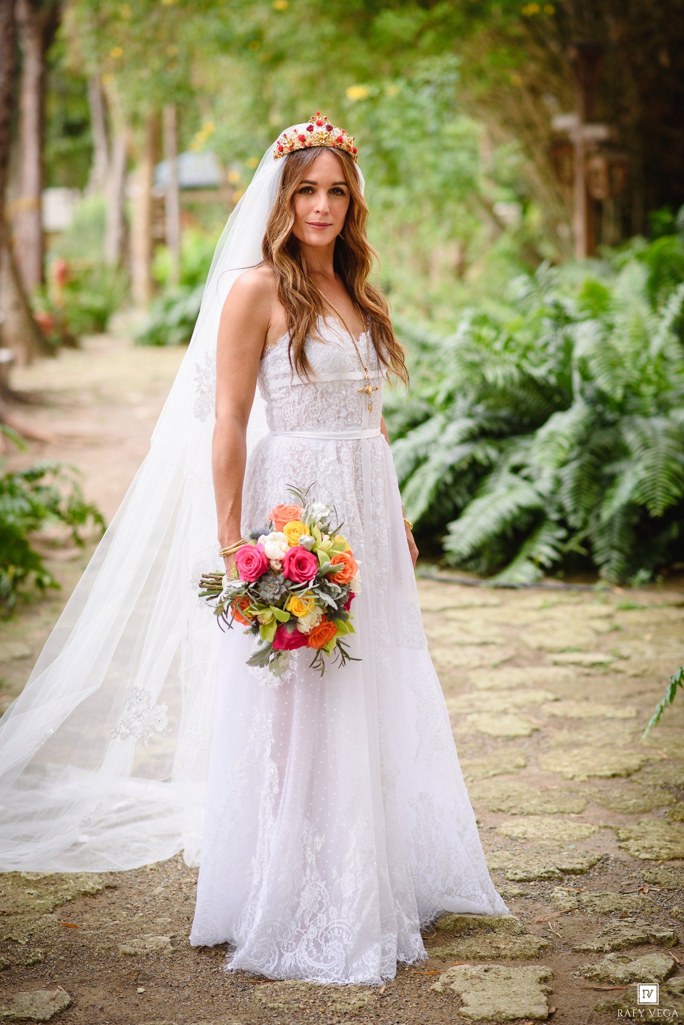 Bride in white lace gown with floral crown and colorful bouquet standing on stone path in lush garden.