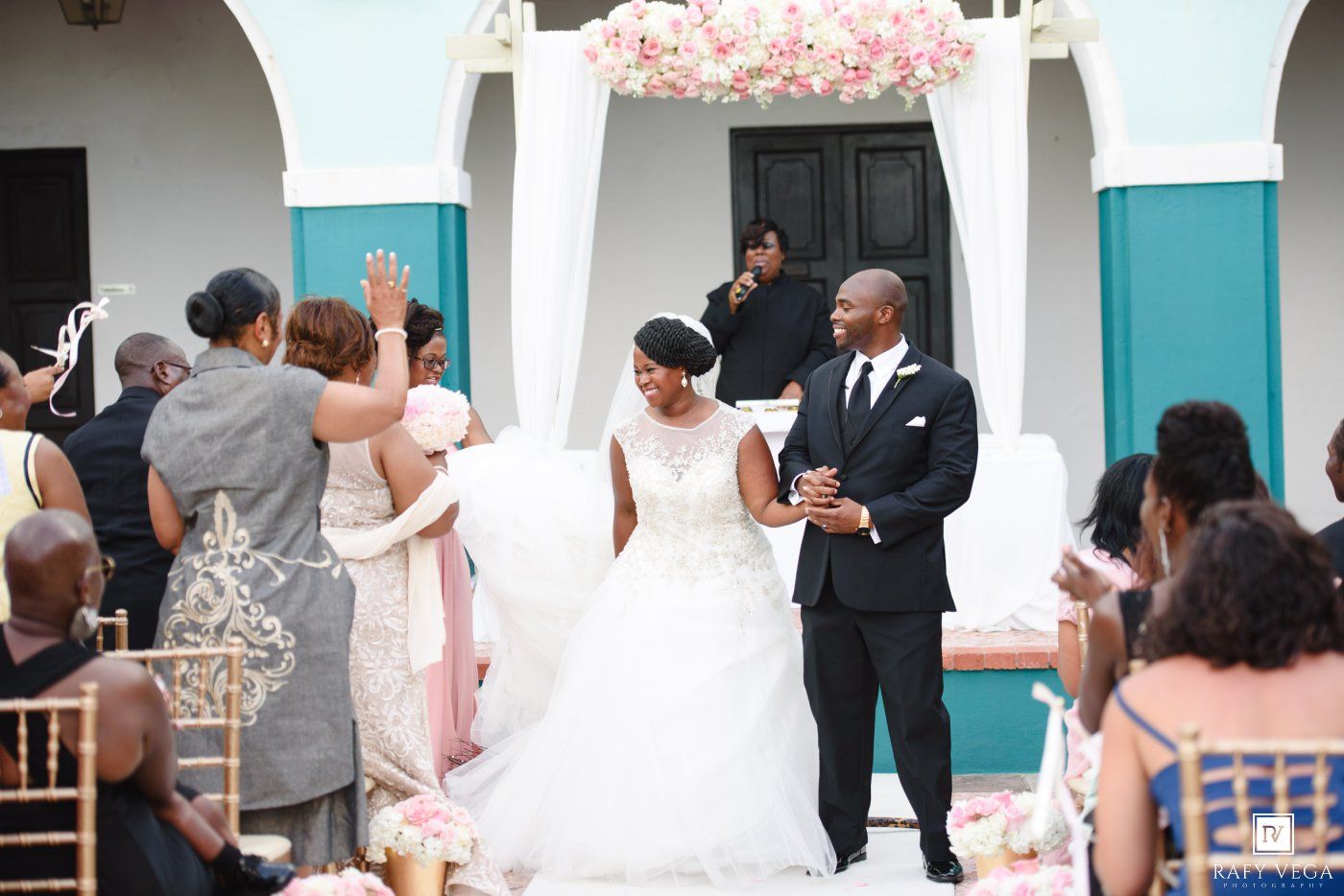 Bride and groom walk down the aisle at an outdoor wedding ceremony with floral arch and guests applauding.