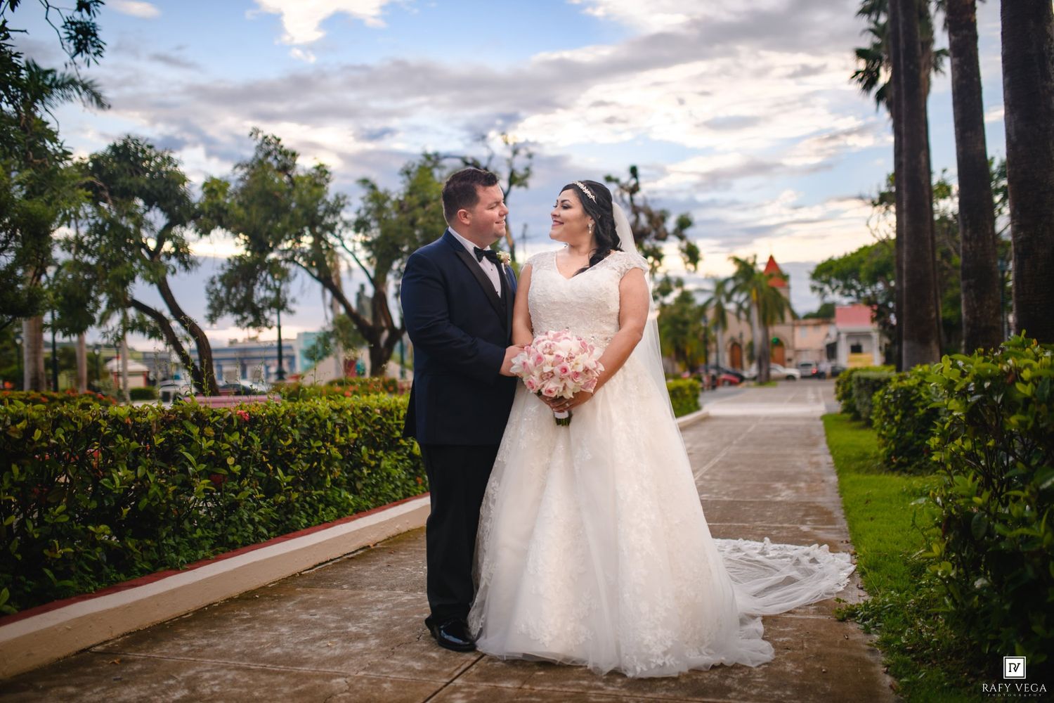 A bride and groom share a romantic moment on a garden pathway, with lush greenery and palm trees in the background.