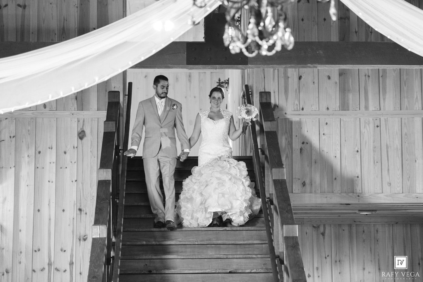 Bride and groom descend wooden staircase in rustic barn venue in elegant black and white wedding photo.