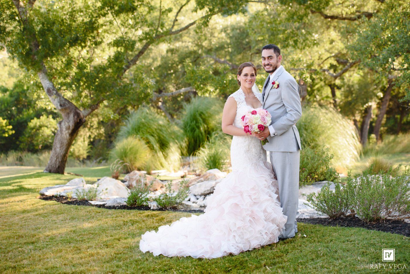Bride in ruffled white gown and groom in gray suit pose together outdoors in a lush garden wedding portrait.