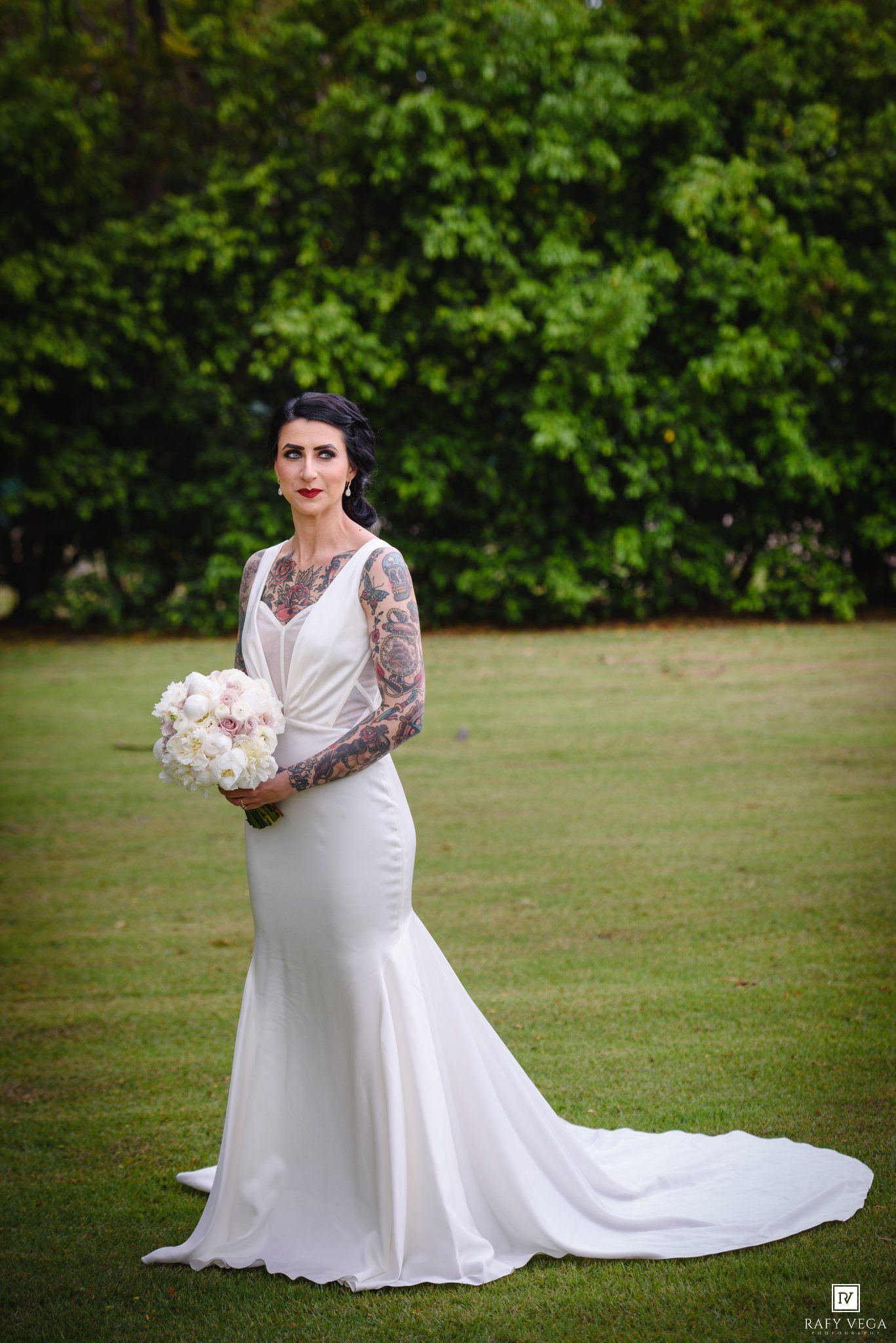 Tattooed bride in fitted white mermaid gown holding bouquet stands on green lawn with lush trees backdrop.