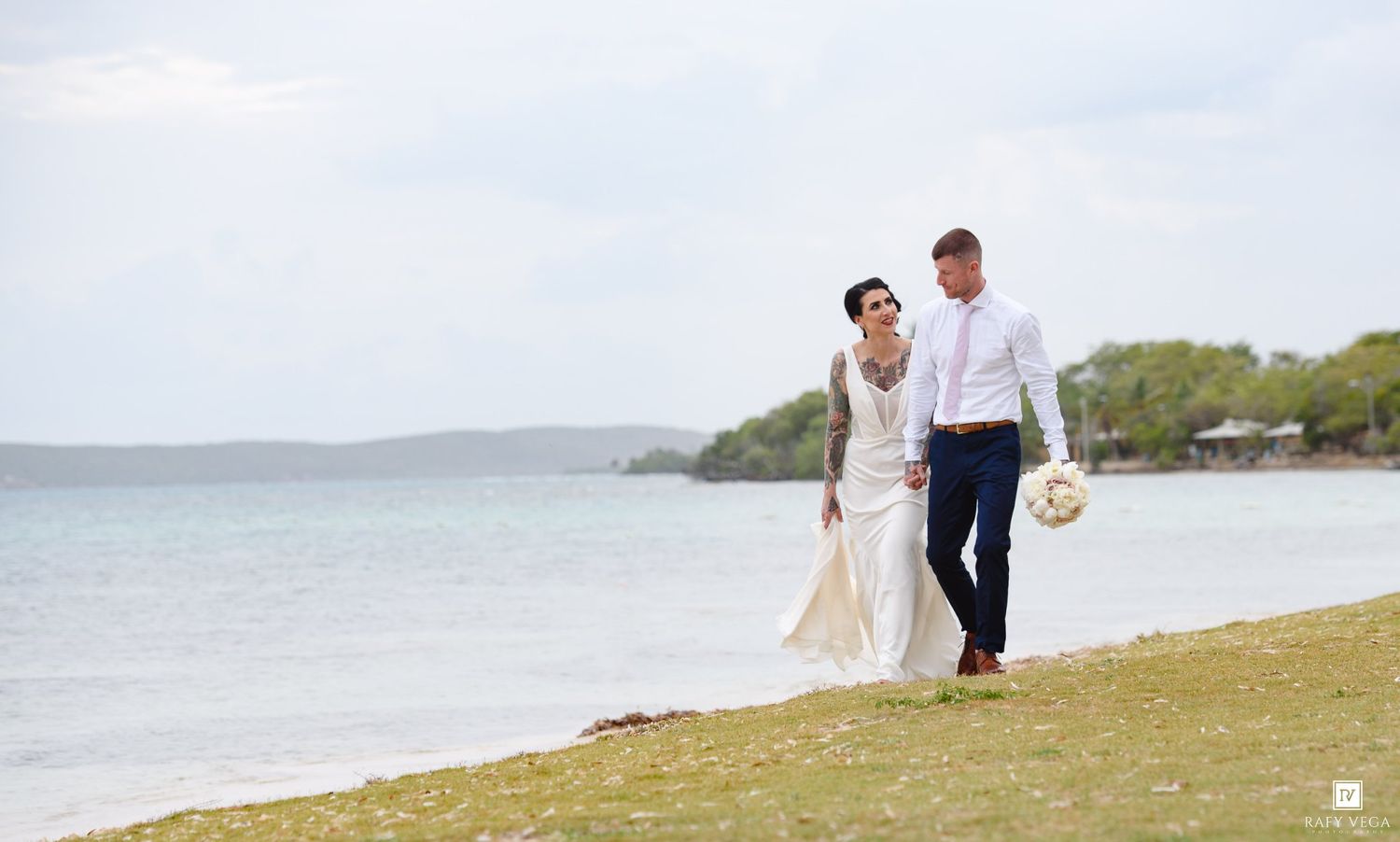 Bride and groom walk hand in hand along a scenic waterfront on their wedding day.