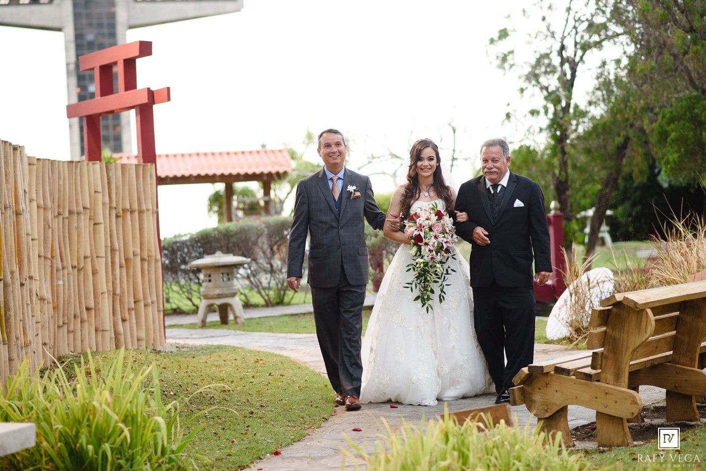 Bride walks down outdoor path flanked by two men in suits, holding pink floral bouquet at a garden wedding.