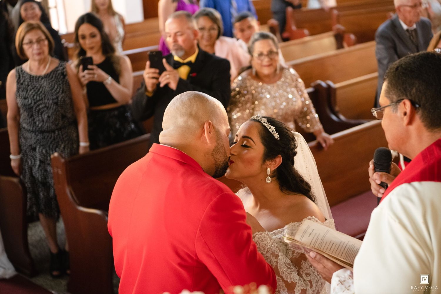 Bride and groom share first kiss at church altar during wedding ceremony with guests watching in pews.