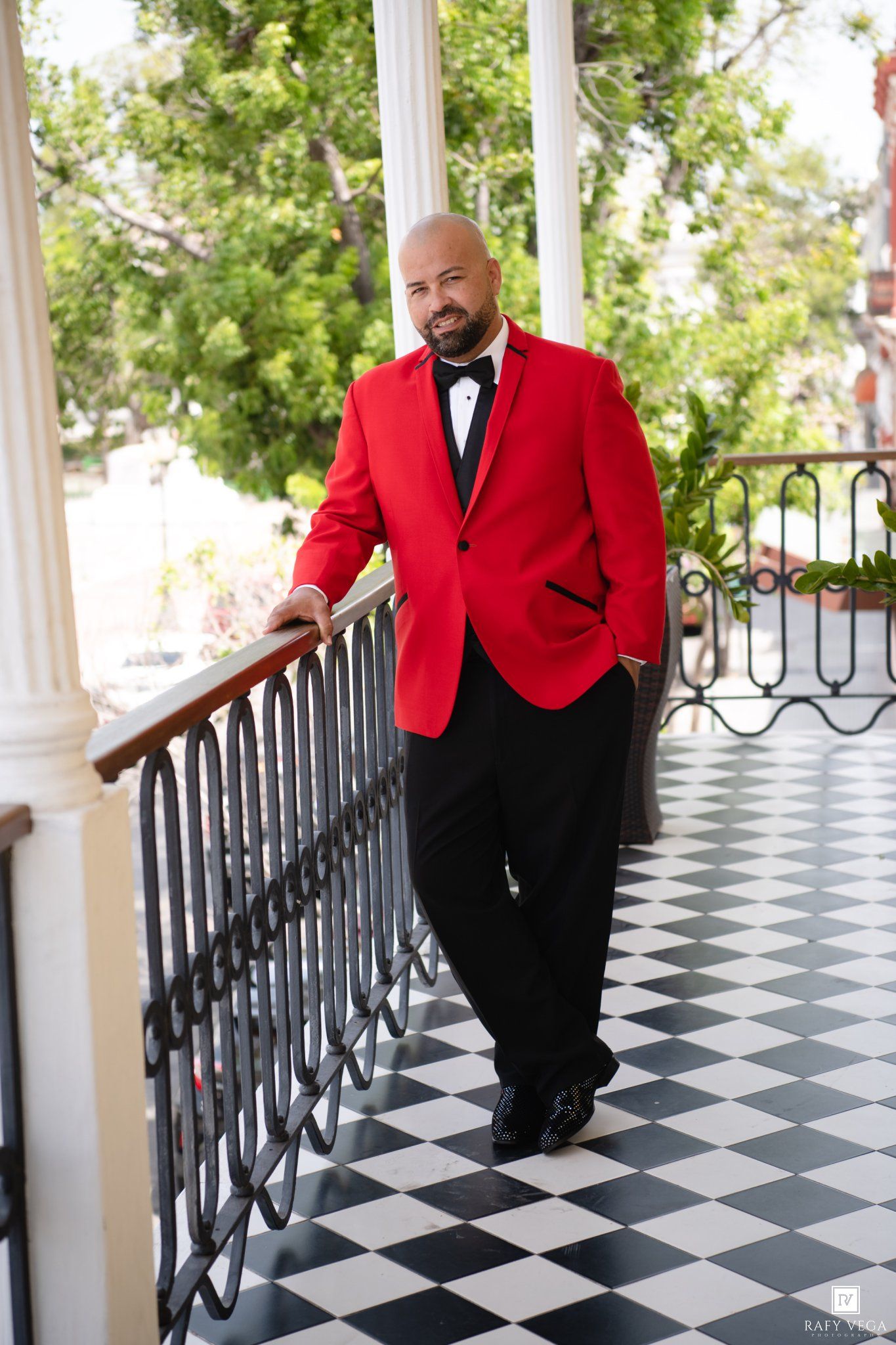 Bald man in red tuxedo jacket and black bow tie leaning on ornate railing of elegant checkered floor porch.