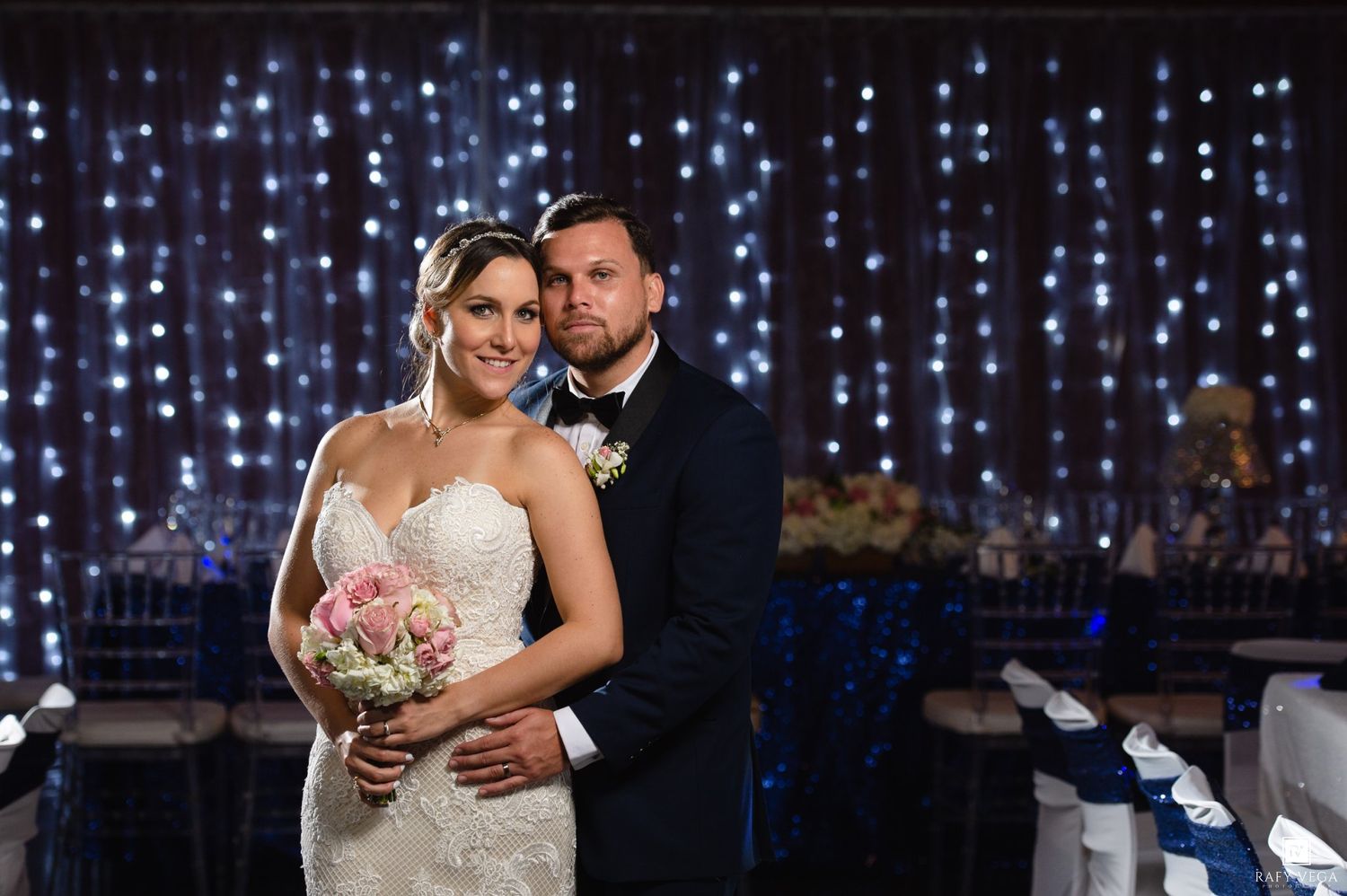 Bride and groom pose elegantly in front of a sparkling fairy light backdrop at their wedding reception.