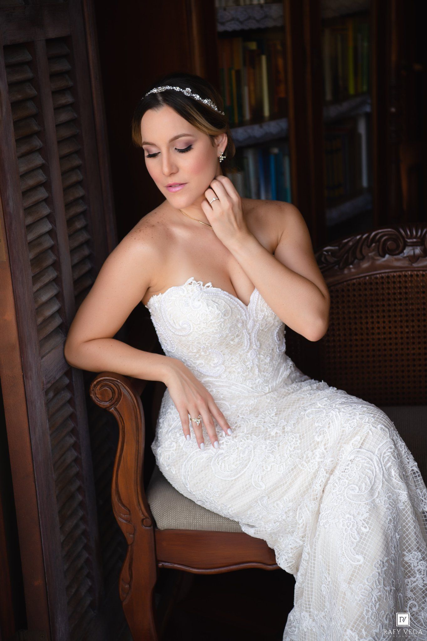 Bride in strapless white lace wedding gown with tiara sits elegantly in ornate chair, touching her earring.