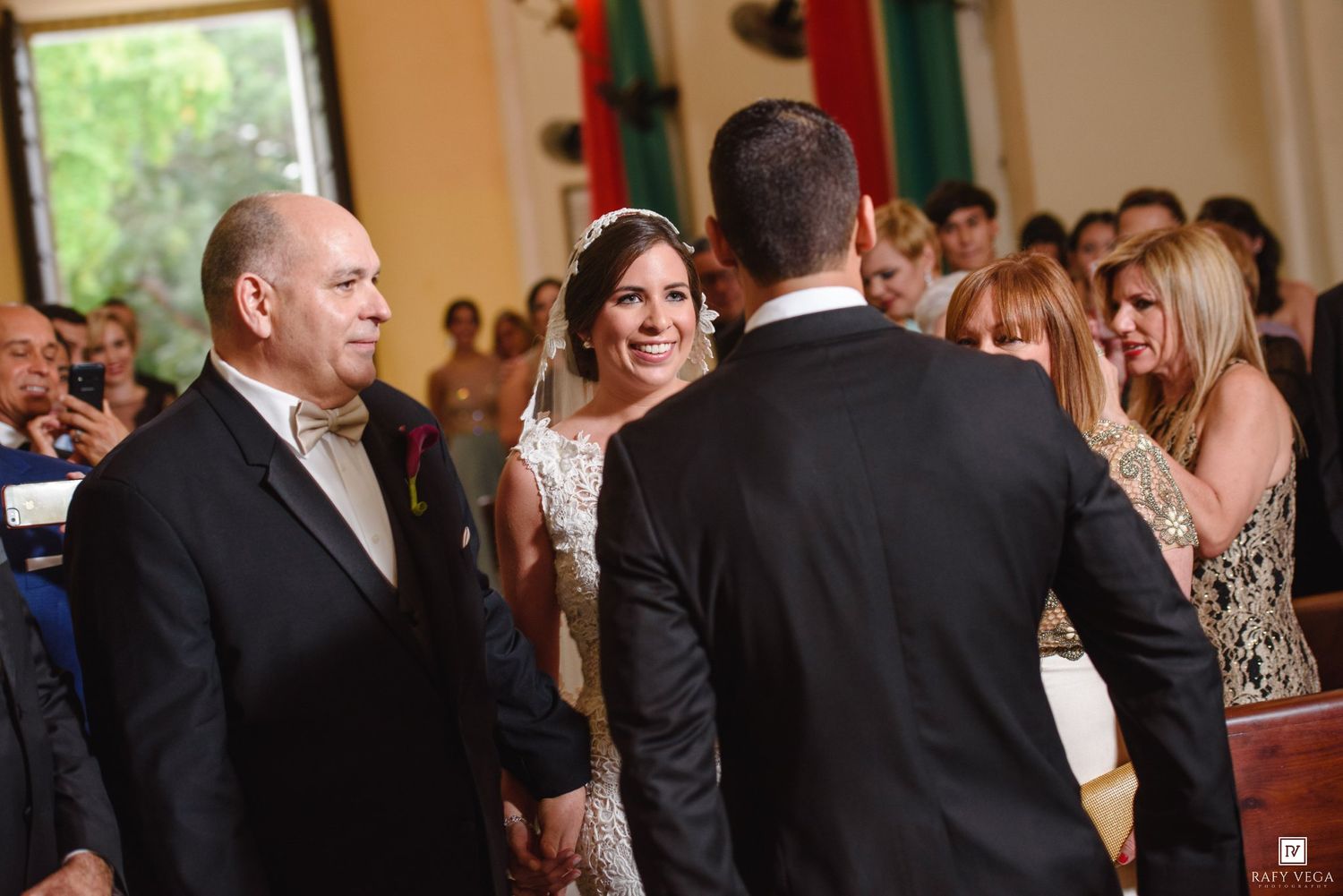 Bride smiling at groom during wedding ceremony while older man stands beside her in elegant church setting.