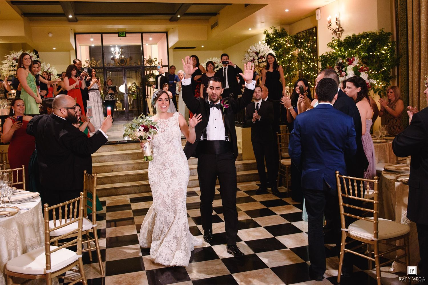 Bride and groom wave joyfully as they enter their elegant wedding reception on a black and white checkered dance floor.