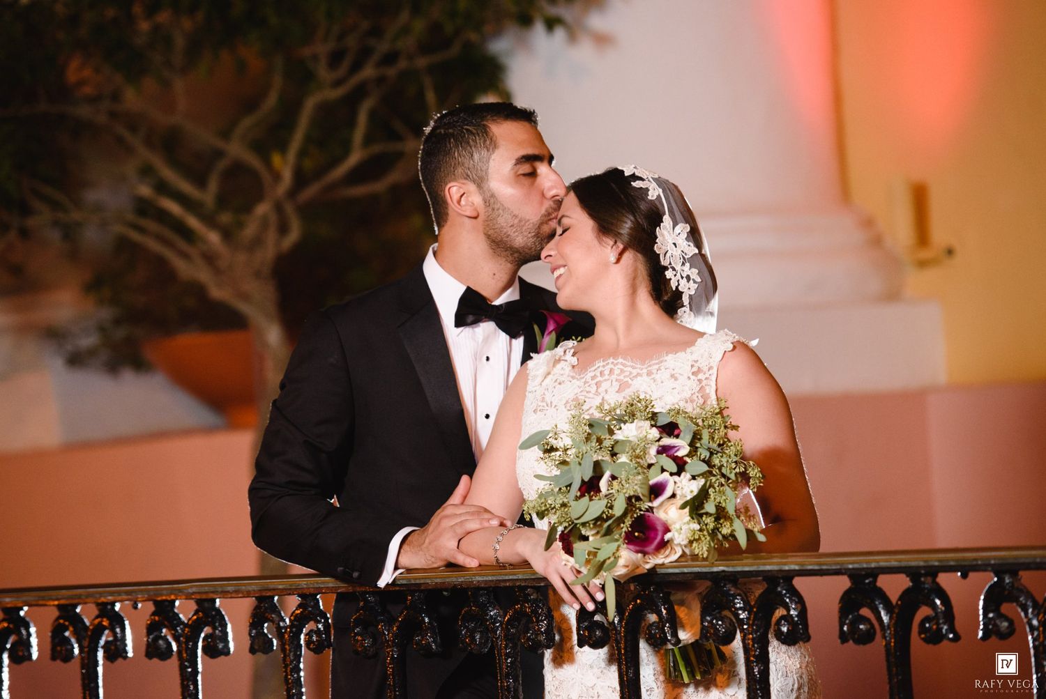 A groom in a tuxedo kisses his bride in a white gown holding a floral bouquet on a balcony at night.