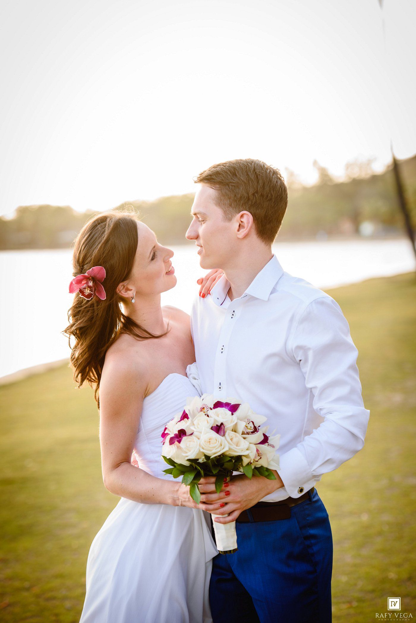 Bride and groom share a romantic moment outdoors near a lake, she holds a white and purple bouquet, wearing a strapless gown.