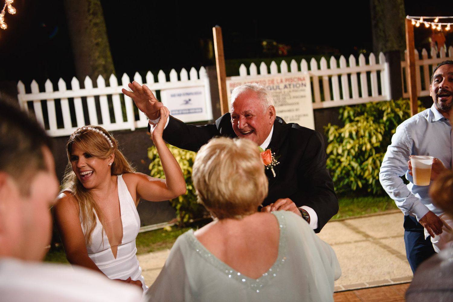 Bride and older man dancing joyfully at outdoor wedding reception at night with white picket fence.