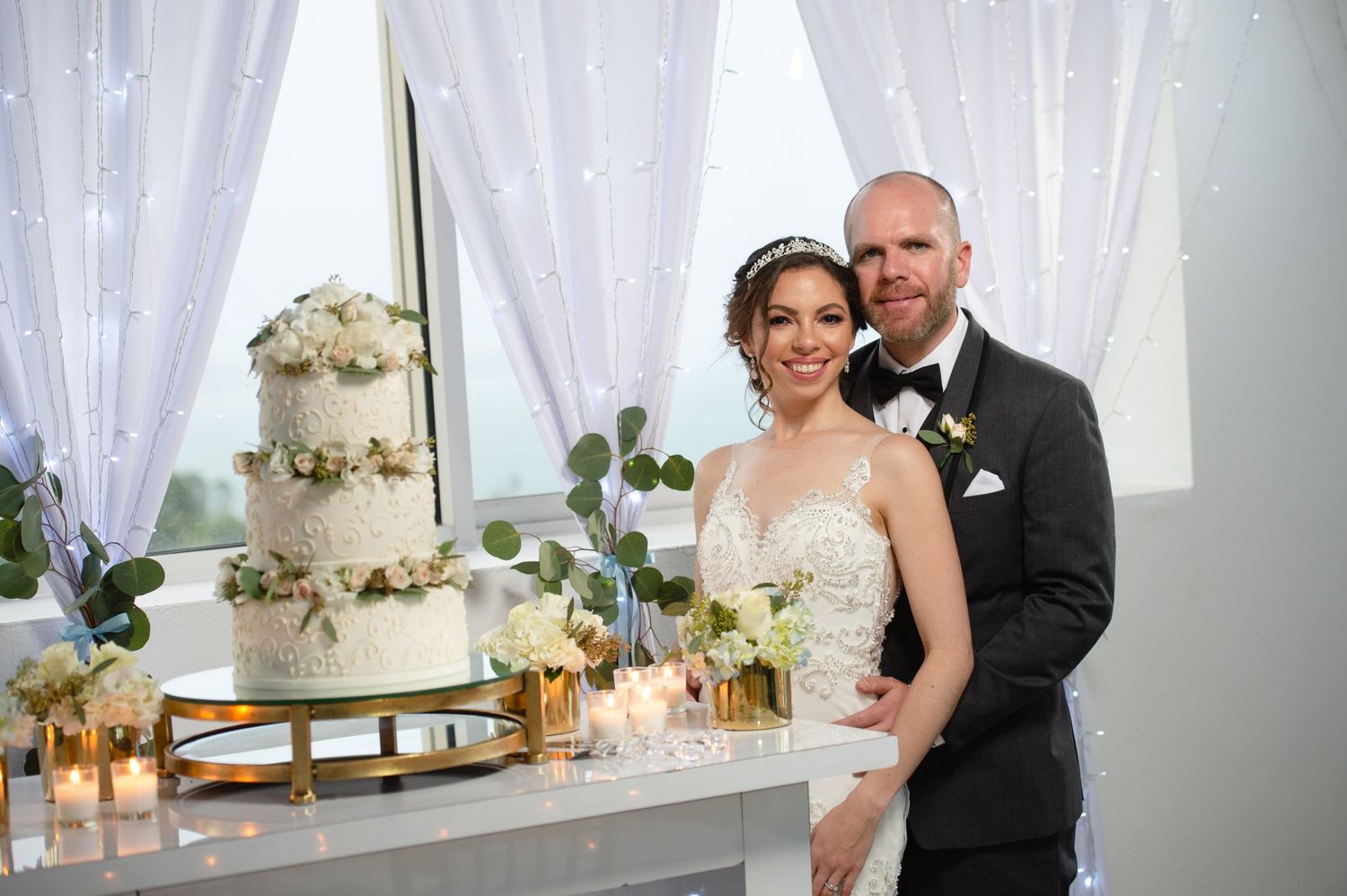 Bride and groom posing together beside a elegant three-tier white floral wedding cake with candles and greenery.