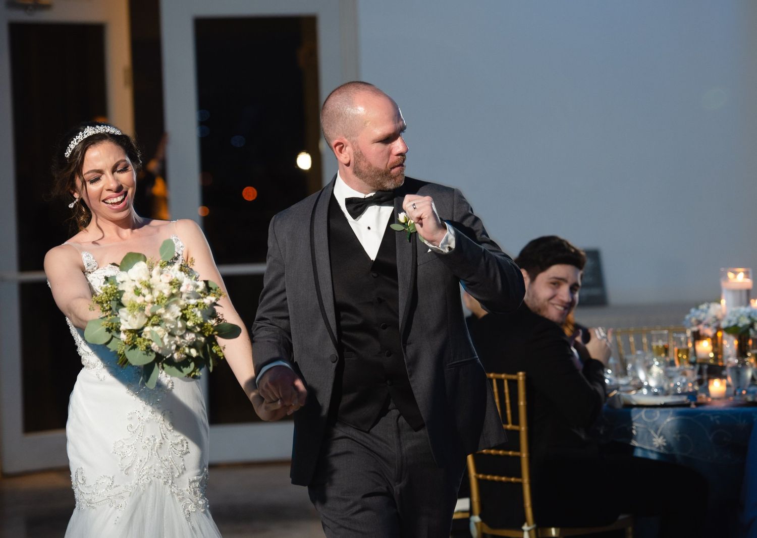 Bride and groom walk hand-in-hand smiling at wedding reception, bride holding white bouquet in elegant venue.