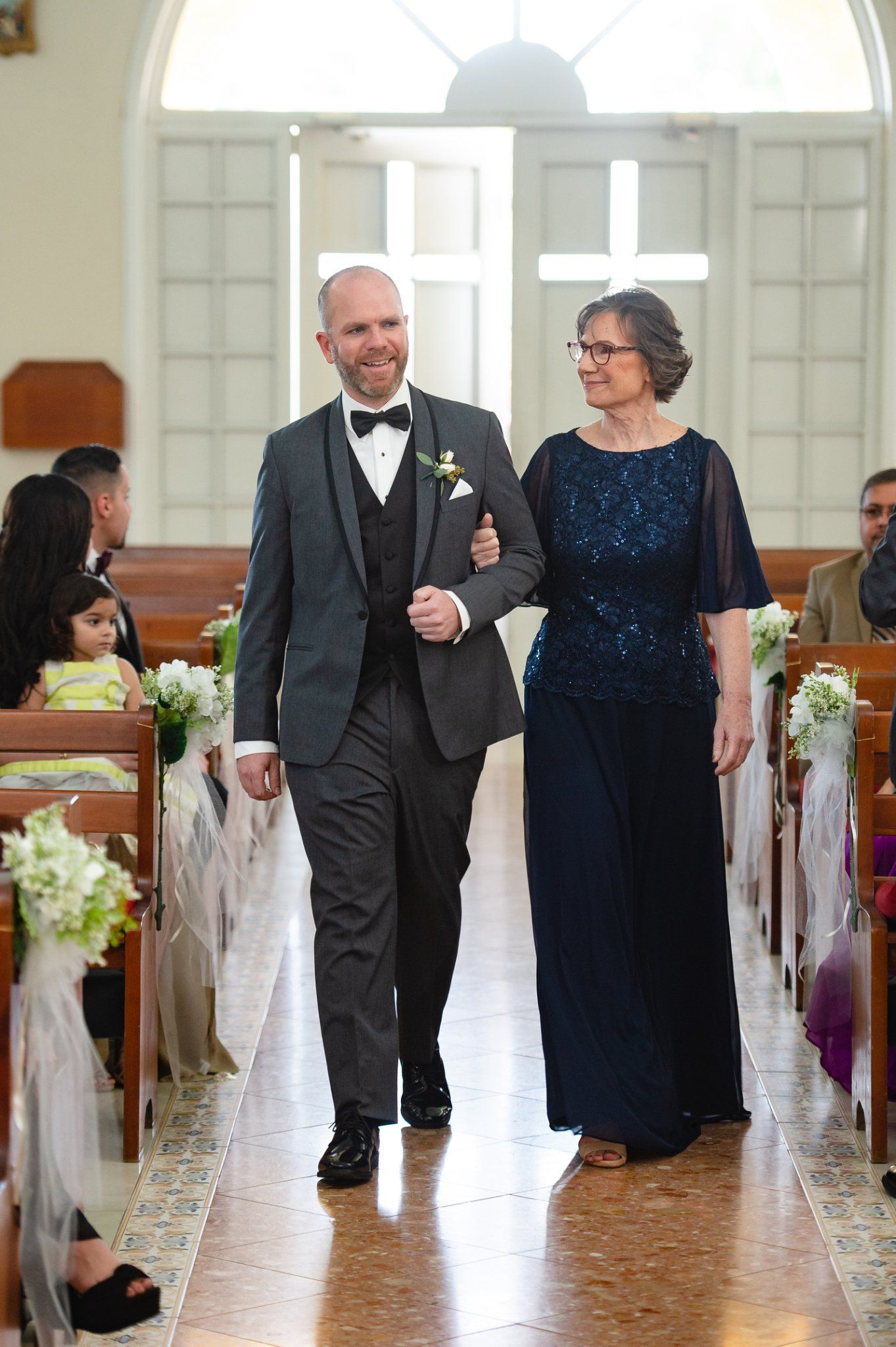Groom in black tuxedo walks down church aisle with older woman in navy blue lace dress during wedding ceremony.