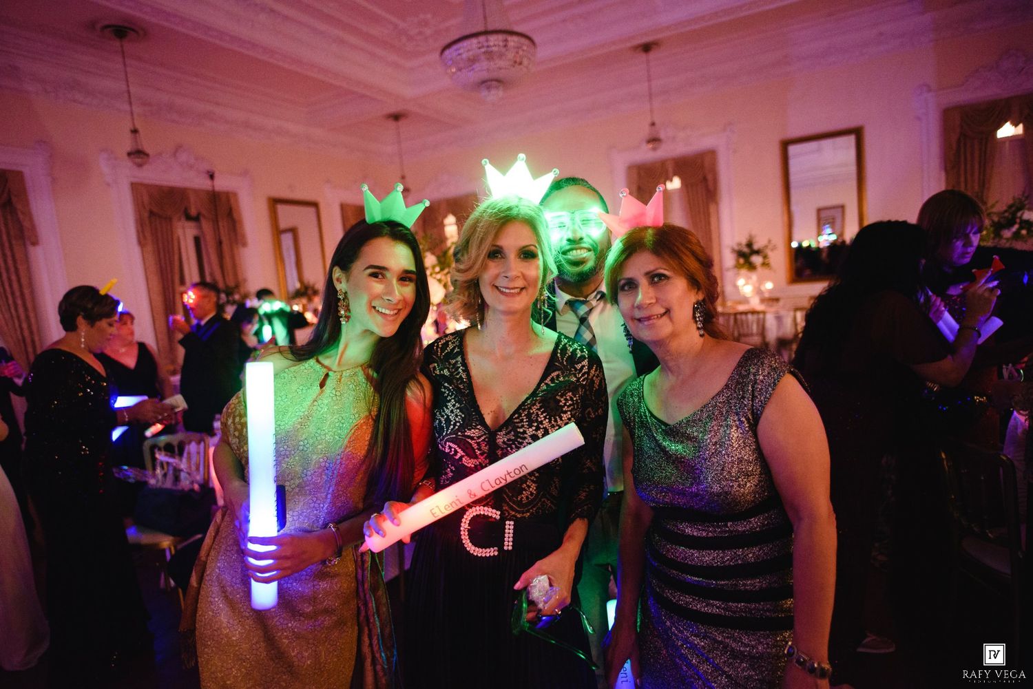 Four women wearing crown headbands pose with colorful glow sticks at a festive wedding.