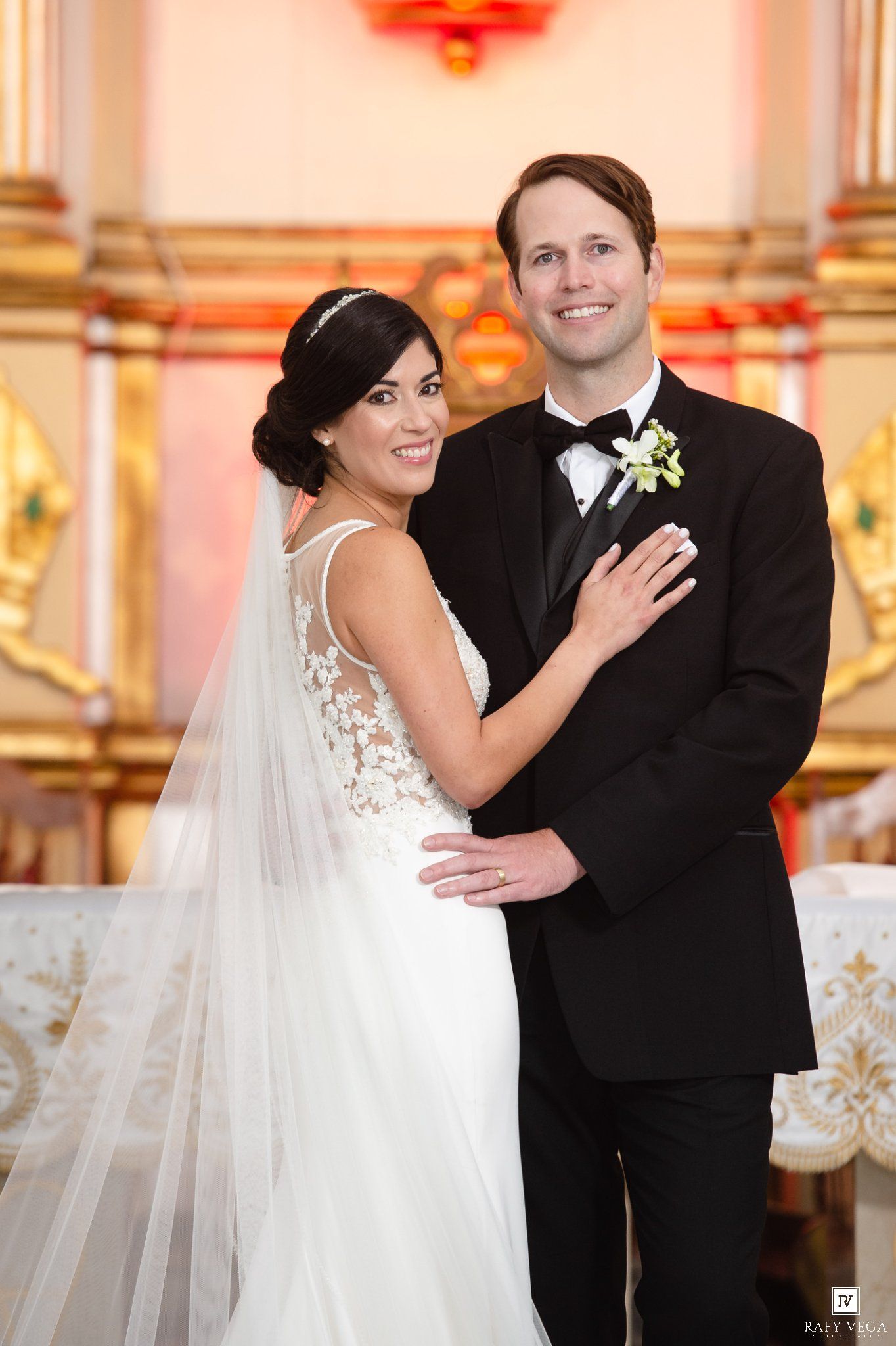 Bride in lace wedding gown and groom in black tuxedo pose together inside an ornate church after their ceremony.