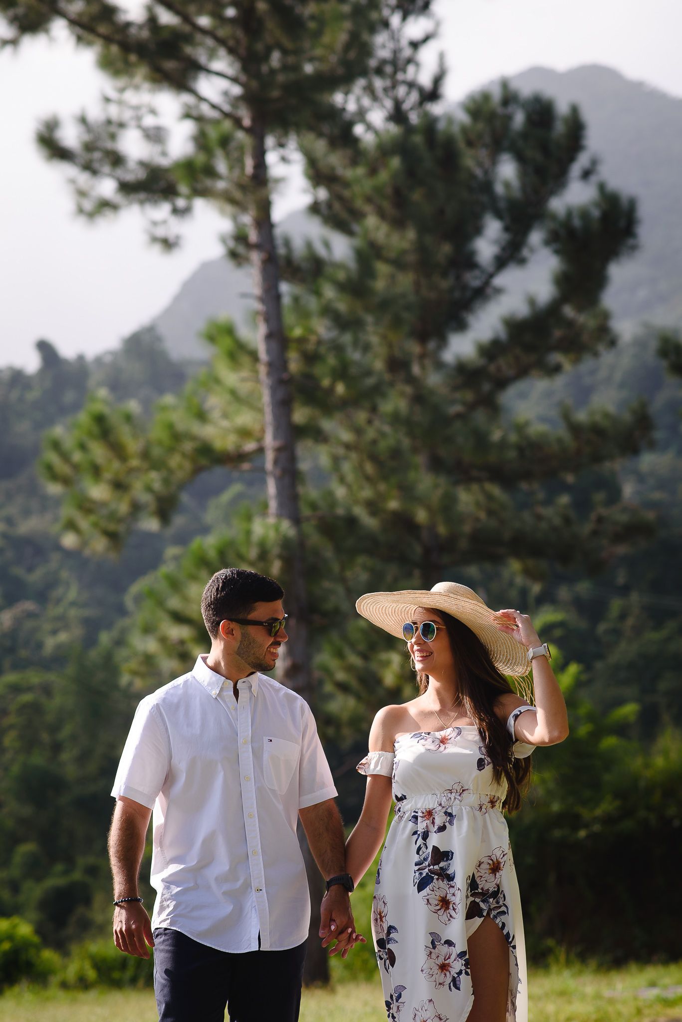 Couple holding hands walking outdoors in nature, woman in floral dress and sun hat, man in white shirt, mountains behind.