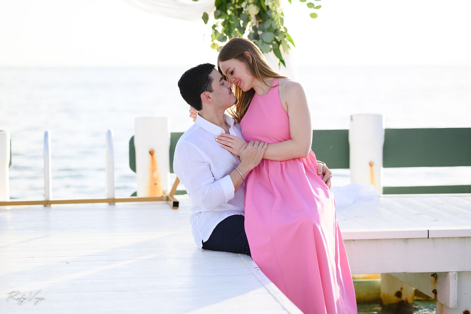 Couple sharing a romantic moment on a sunlit dock, woman in pink dress and man in white shirt near the water.